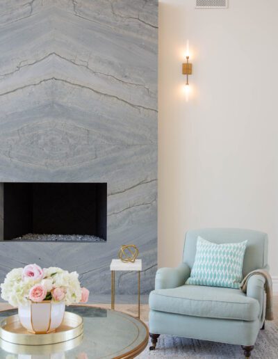 Elegant living room corner with a chevron-patterned marble fireplace, soft blue armchair, gold-accented side table, and a decorative wall sconce.