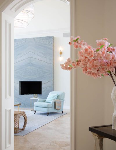 Elegant interior view through an arched doorway featuring a living room with marble fireplace, pastel armchair, and floral decor.