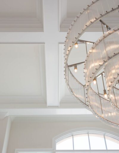 Elegant chandelier with circular design hanging from a white ceiling with crown molding, near an arched window streaming in natural light.