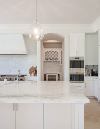 A modern kitchen interior featuring white cabinetry, marble countertops, stainless steel appliances, and a pendant light.