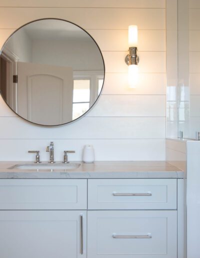 A modern bathroom interior featuring a round mirror above a white vanity with a marble countertop and a wall-mounted sconce to the side.