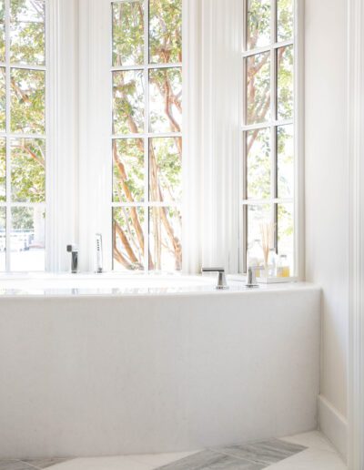 A bright, minimalist bathroom with a large white tub in front of tall windows showing trees outside.