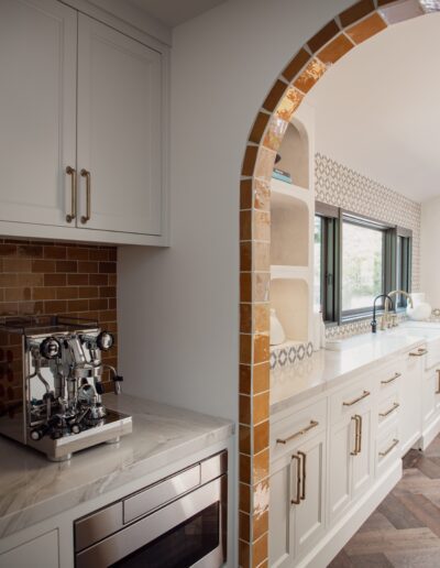 A modern kitchen featuring a coffee machine, marble countertops, and white cabinets. The room includes an archway with orange tiles and a large window above the sink letting in natural light.