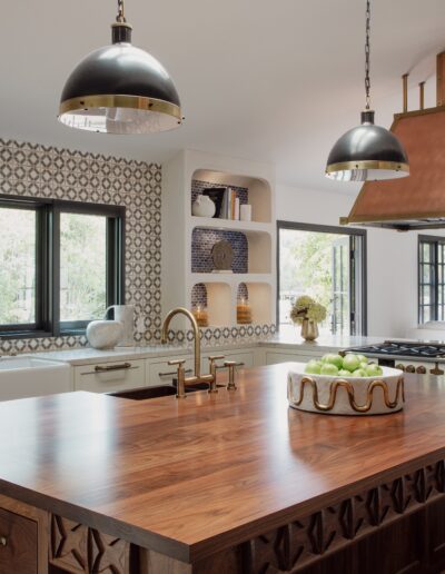 A modern kitchen with a wooden island, patterned backsplash, copper range hood, and two pendant lights. Shelves with decor items are visible, and the windows offer a view of trees outside.