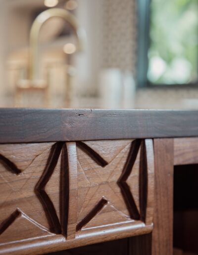 Close-up of a wooden kitchen island edge with intricate carved patterns, and a blurry background showing a sink with a brass faucet and a window.