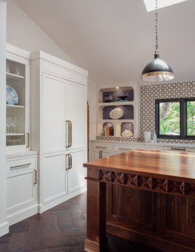 A kitchen with white cabinetry, a wooden island, patterned backsplash, and dark wood flooring. A dome-shaped pendant light hangs from the ceiling, and a window lets in natural light.