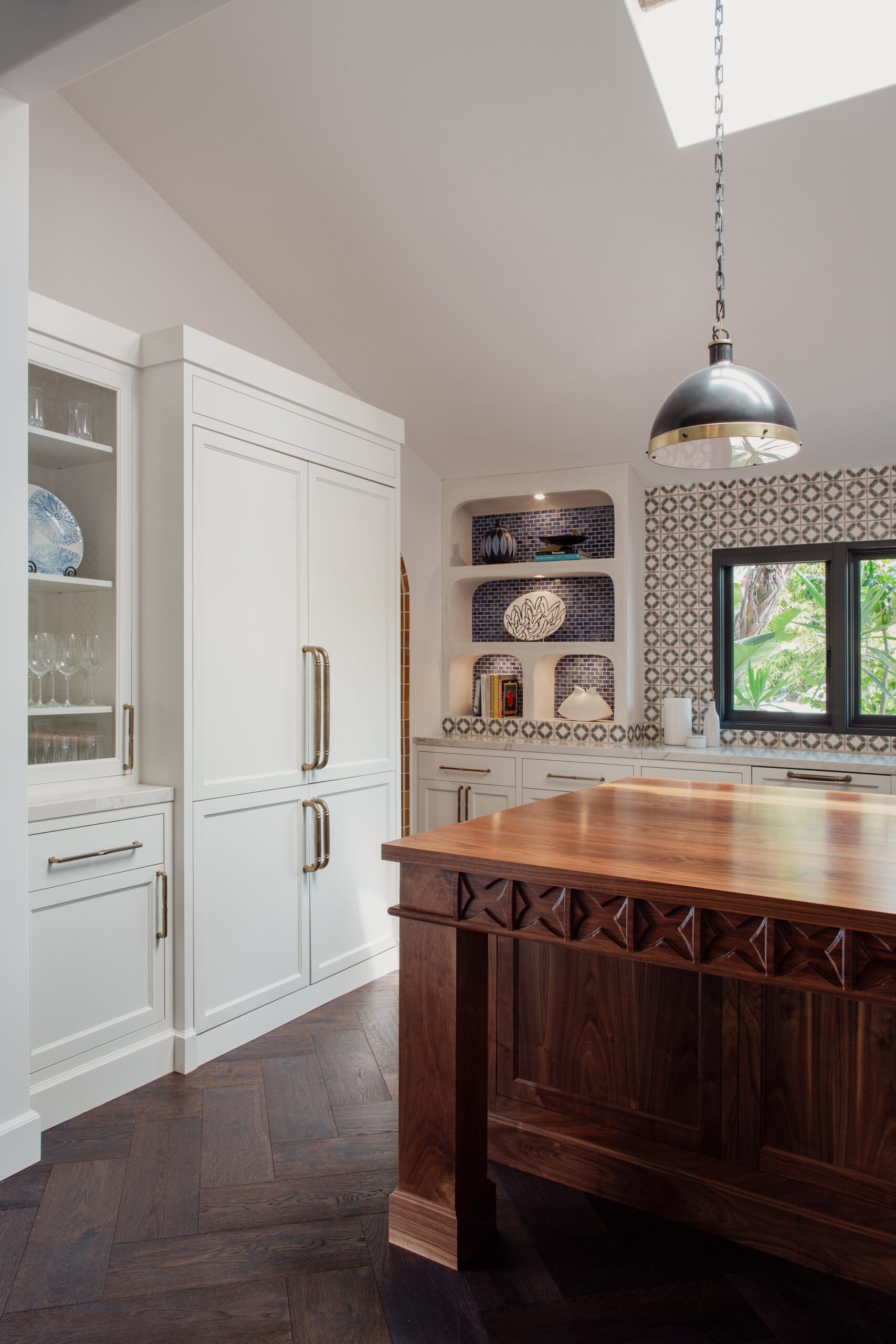 A kitchen with white cabinetry, a wooden island, patterned backsplash, and dark wood flooring. A dome-shaped pendant light hangs from the ceiling, and a window lets in natural light.