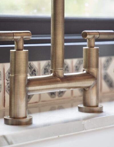 A brass kitchen faucet and matching sprayer are installed on a marble countertop with patterned tiles in the background.