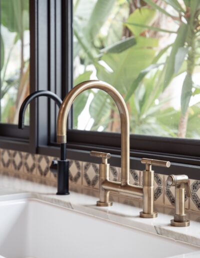 Close-up of a kitchen sink with a brass faucet and a black lever. A large window with a view of lush green plants is in the background. Patterned tiles adorn the wall behind the sink.