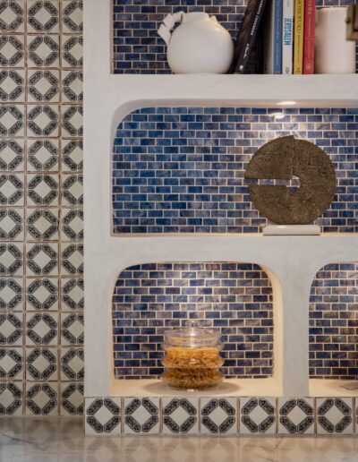 A white shelf with blue mosaic tiles holds a few books, a white teapot, a circular sculpture, and a glass jar filled with cereal. The wall has a black and white patterned tile design.