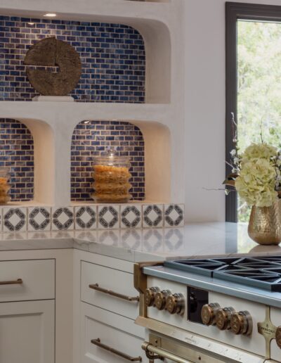 A kitchen with white cabinets, a gas stove, and a decorative blue tile backsplash. Shelves display candles in front of blue mosaic tiles, and a vase with flowers is on the counter. A window shows greenery outside.