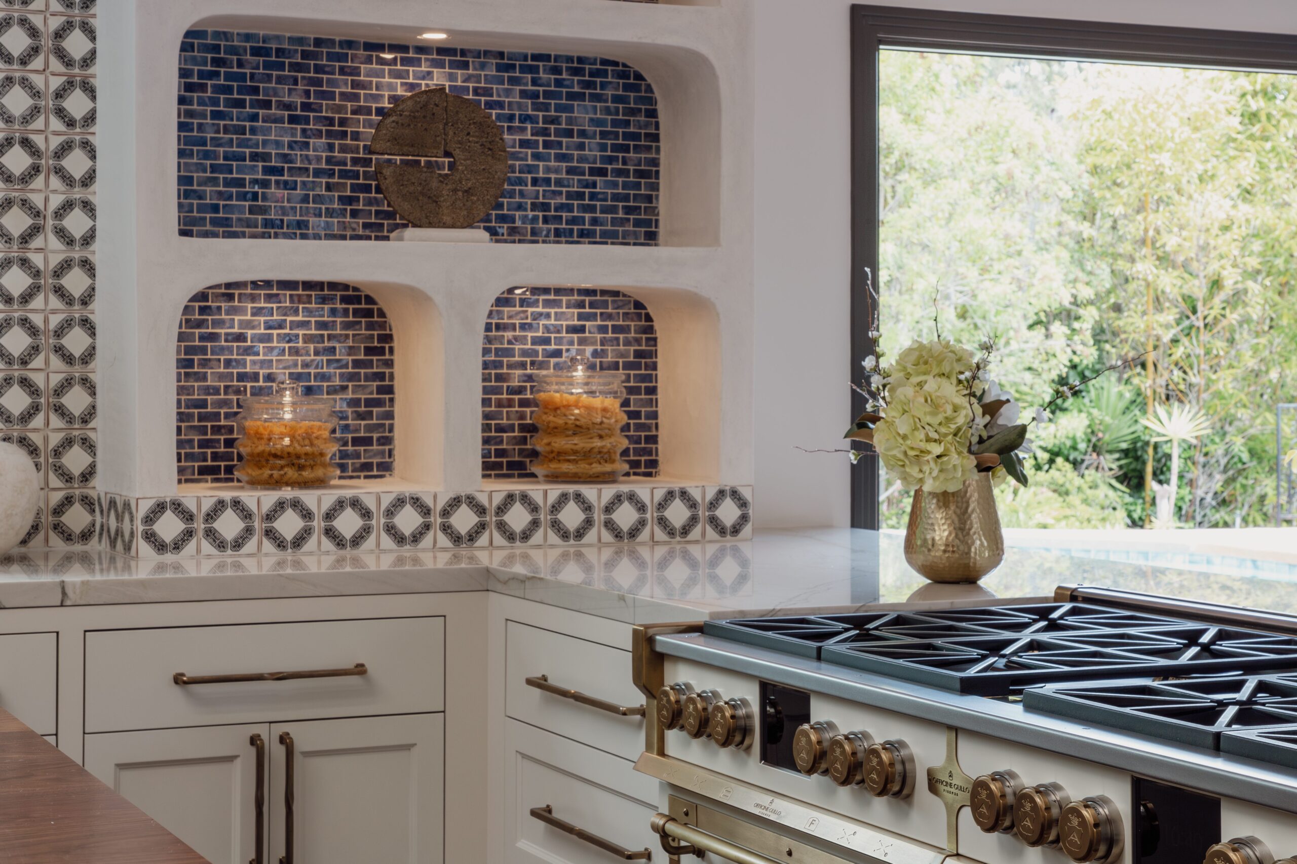 A kitchen with white cabinets, a gas stove, and a decorative blue tile backsplash. Shelves display candles in front of blue mosaic tiles, and a vase with flowers is on the counter. A window shows greenery outside.