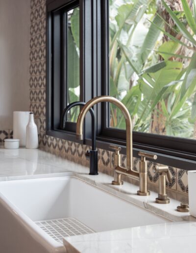 A modern kitchen sink with brass faucets, a white countertop, and patterned backsplash, set against a large window overlooking green plants.