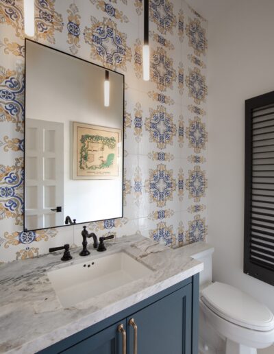 A bathroom with decorative blue and gold tile backsplash, a rectangular mirror, a marble countertop, a sink with black fixtures, a blue vanity, and a toilet.