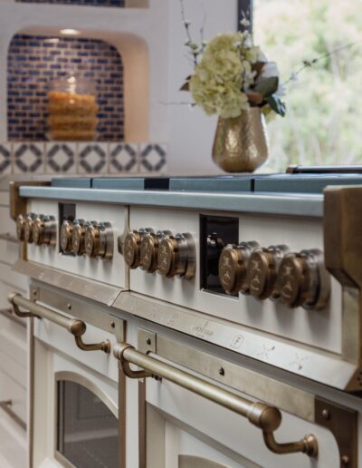 A close-up view of a modern kitchen stove with brass handles and knobs. The backsplash features blue tiles and two jars placed in niches. A vase with flowers sits on the counter.