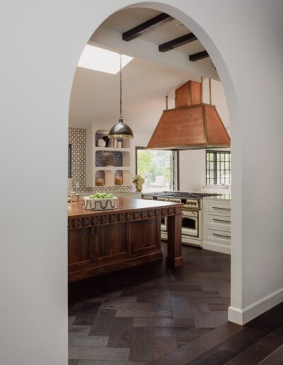 View through an arched doorway of a modern kitchen with dark wood flooring, a large copper exhaust hood, a wooden island, white cabinets, and patterned backsplash tiles.