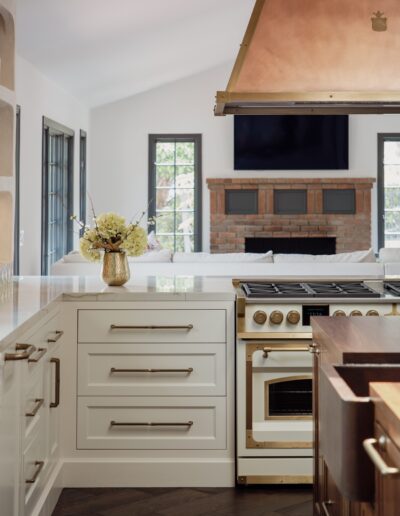 A modern kitchen with white cabinetry, a gas stove with brass accents, and a white countertop with a vase of flowers. A brick fireplace and large windows are visible in the background.