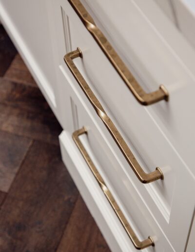 Close-up of three white cabinet drawers with long, gold handles, set against a dark wood floor.