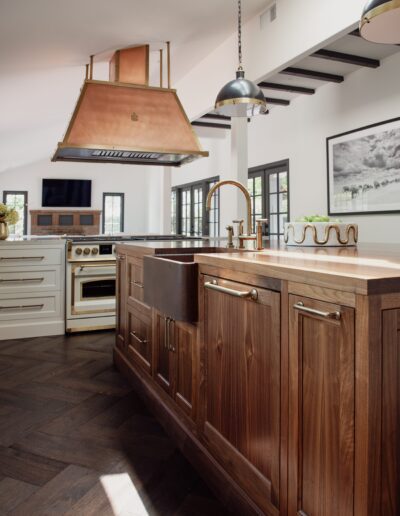 Modern kitchen with a wooden island, copper range hood, farmhouse sink, and built-in oven. The kitchen features stainless steel fixtures, pendant lighting, wooden flooring, and a lighted ceiling.