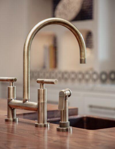 Close-up of a modern kitchen sink with a brass faucet and sprayer. The countertop is wooden, and part of the kitchen's backsplash is visible in the background.