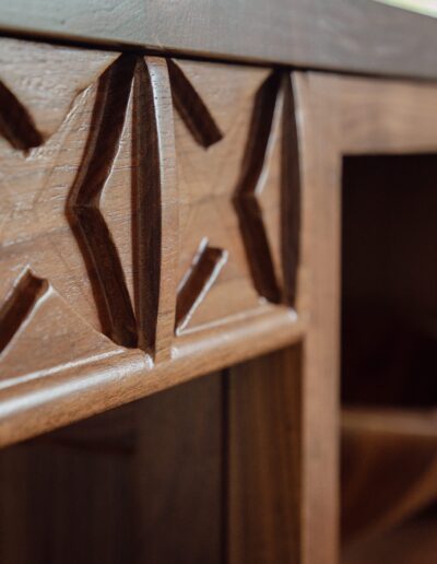 A close-up of a wooden cabinet featuring intricate carved X patterns on the front panel and an open shelf with a wooden bowl inside.