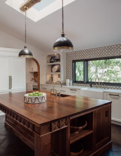 A modern kitchen featuring a large wooden island with a sink, two hanging black pendant lights, white cabinets, patterned backsplash, and a skylight above. There are decorative items and fruit on the island.