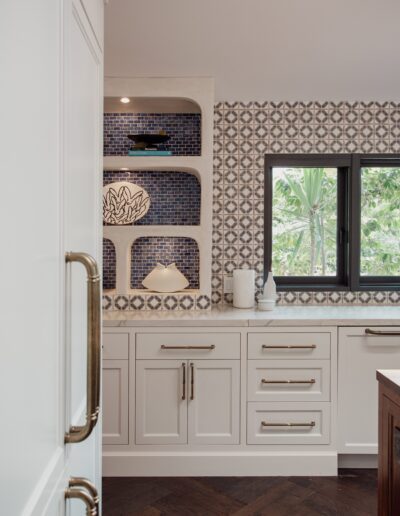 A kitchen with white cabinets, brass handles, and a patterned tile backsplash. Open shelves with decor, a window, and a dark wood floor are also visible.