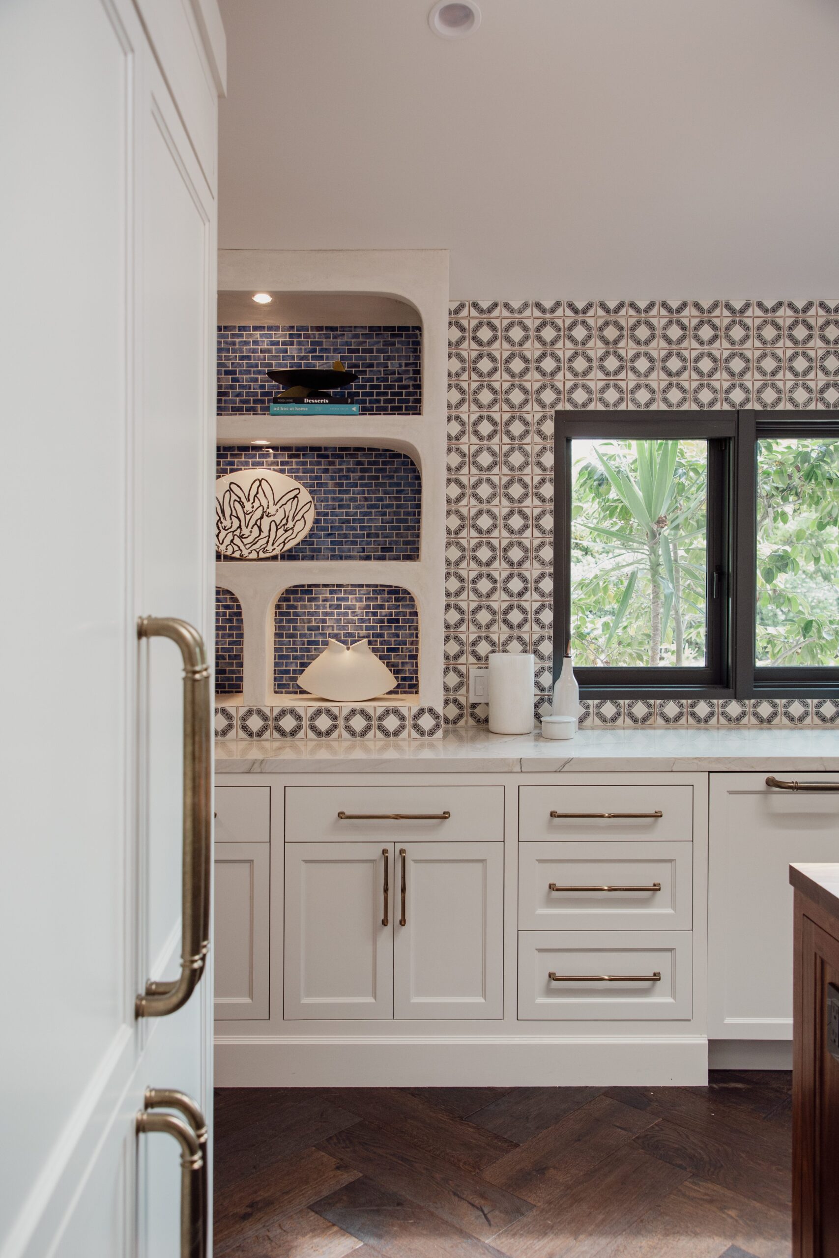 A kitchen with white cabinets, brass handles, and a patterned tile backsplash. Open shelves with decor, a window, and a dark wood floor are also visible.